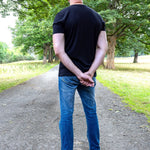 A tall man standing on a dirt path in a park, viewed from behind, wearing a black t-shirt, blue jeans, and gray sneakers.