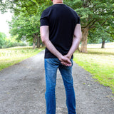 A tall man standing on a dirt path in a park, viewed from behind, wearing a black t-shirt, blue jeans, and gray sneakers.