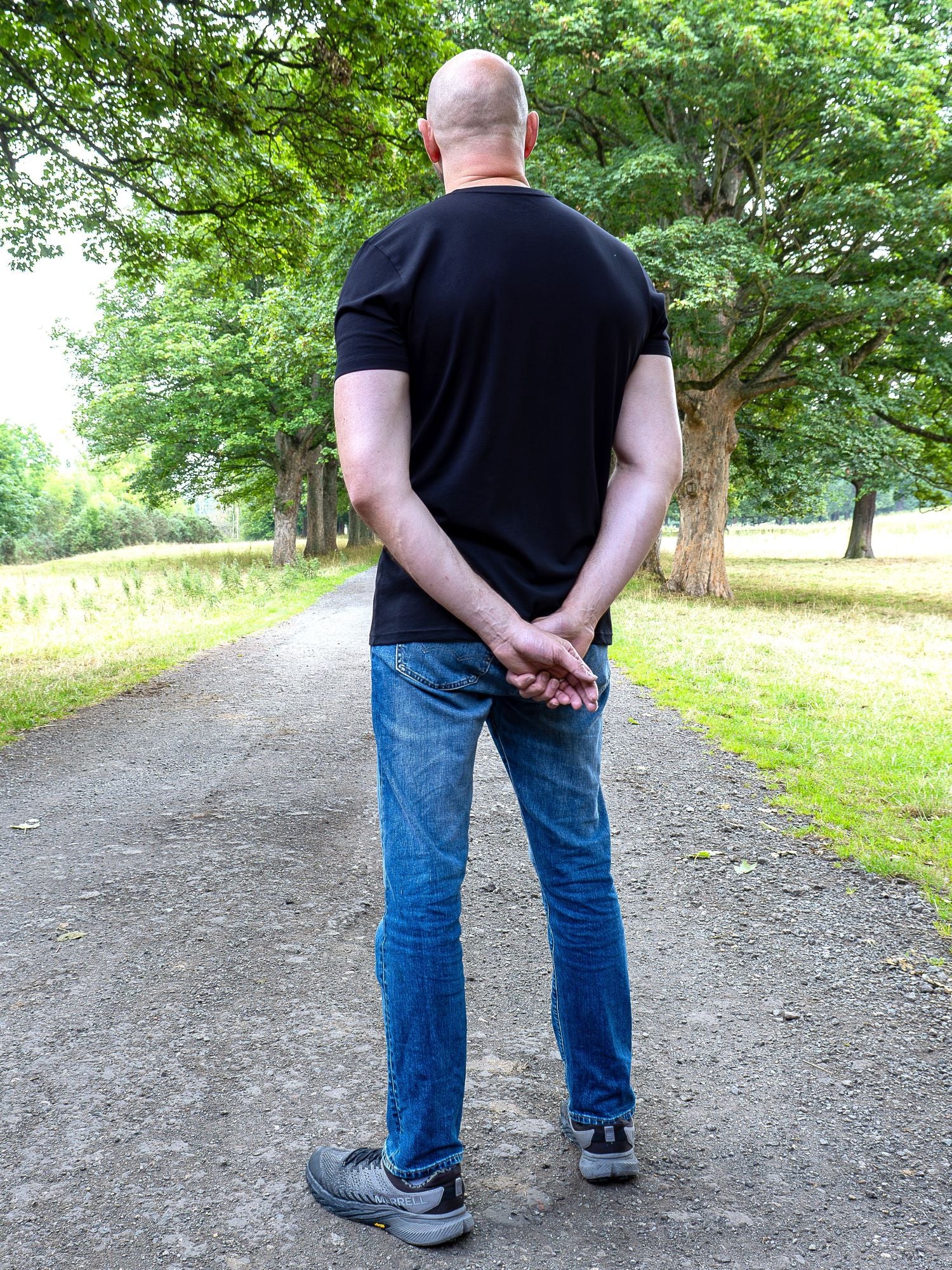 A tall man standing on a dirt path in a park, viewed from behind, wearing a black t-shirt, blue jeans, and gray sneakers.