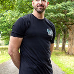 A tall bald man with a beard wearing a black t-shirt with a small white and blue logo on the left chest, standing on a park path with trees in the background.