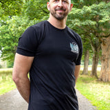 A tall bald man with a beard wearing a black t-shirt with a small white and blue logo on the left chest, standing on a park path with trees in the background.