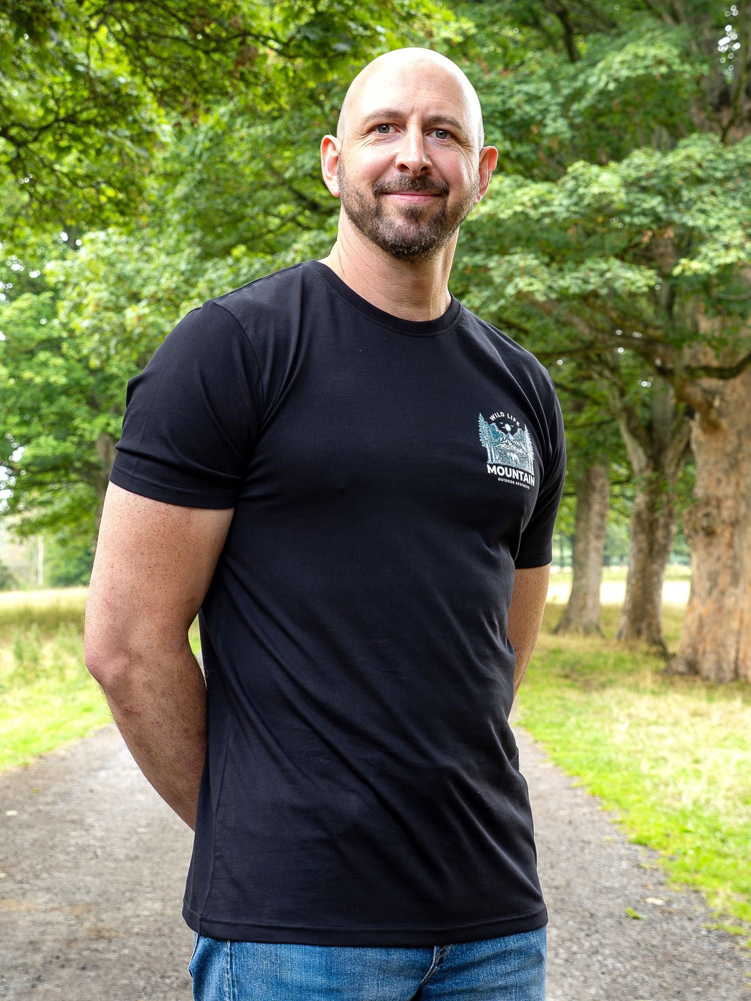 A tall bald man with a beard wearing a black t-shirt with a small white and blue logo on the left chest, standing on a park path with trees in the background.