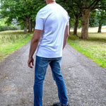 A tall man viewed from behind wearing a plain white short-sleeve t-shirt, blue jeans, and gray sneakers, standing on a gravel path in a park with green trees.