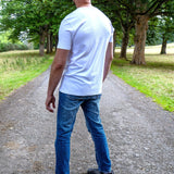 A tall man viewed from behind wearing a plain white short-sleeve t-shirt, blue jeans, and gray sneakers, standing on a gravel path in a park with green trees.