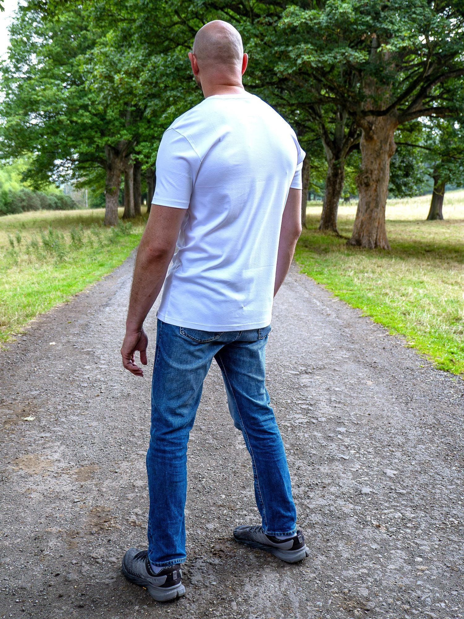A tall man viewed from behind wearing a plain white short-sleeve t-shirt, blue jeans, and gray sneakers, standing on a gravel path in a park with green trees.