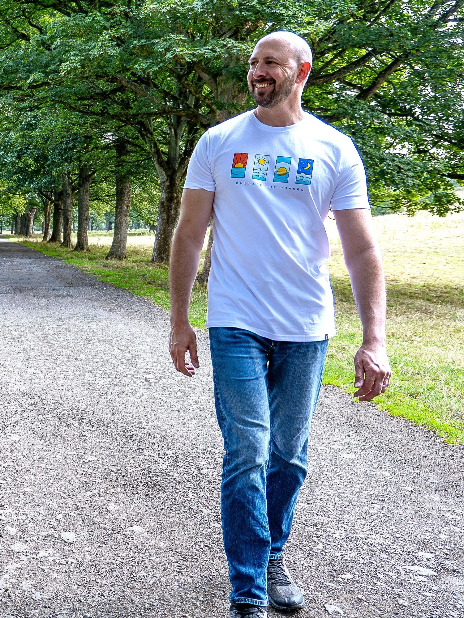 A tall man walking on a gravel path in a park, wearing a white t-shirt with a colorful graphic print and blue jeans.