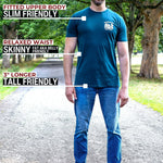 A tall man standing on a tree-lined path wearing a dark teal fitted t-shirt and blue jeans, with text callouts describing the shirt's fit: slim in the upper body, relaxed at the waist, and 3" longer overall.