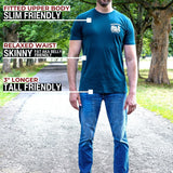 A tall man standing on a tree-lined path wearing a dark teal fitted t-shirt and blue jeans, with text callouts describing the shirt's fit: slim in the upper body, relaxed at the waist, and 3" longer overall.