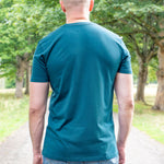 A tall man viewed from behind wearing a teal short-sleeve t-shirt and blue jeans, standing on a gravel path surrounded by green trees.