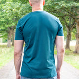 A tall man viewed from behind wearing a teal short-sleeve t-shirt and blue jeans, standing on a gravel path surrounded by green trees.
