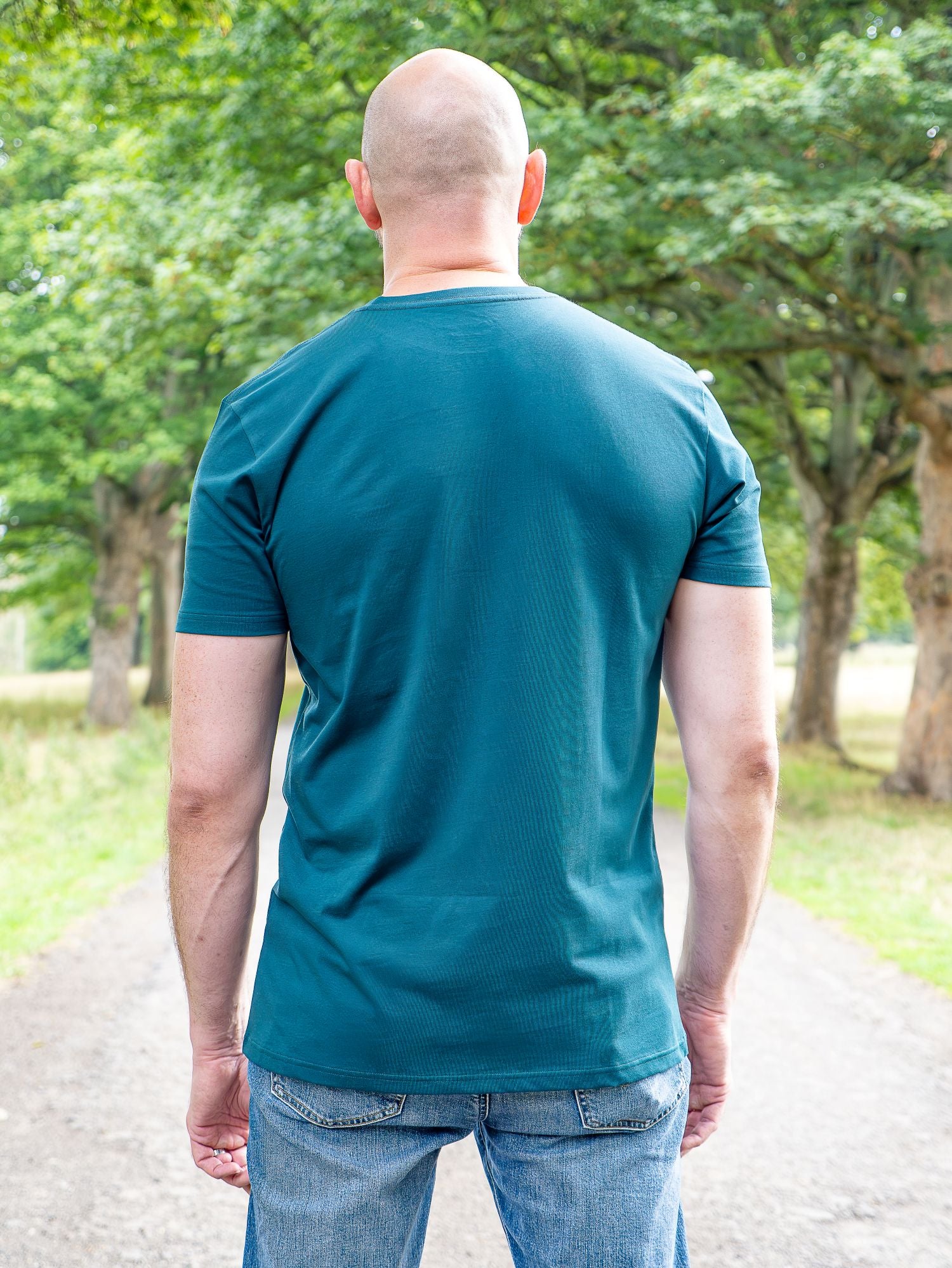 A tall man viewed from behind wearing a teal short-sleeve t-shirt and blue jeans, standing on a gravel path surrounded by green trees.