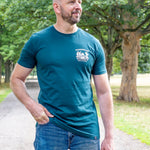 A tall man wearing a teal blue t-shirt with a white crest logo on the left chest, paired with blue jeans, standing outdoors on a park path with trees in the background.