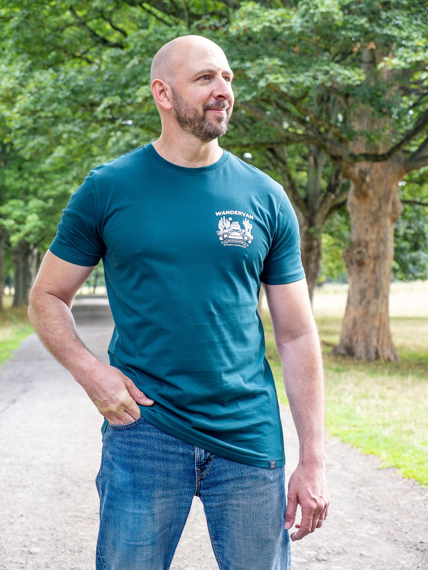 A tall man wearing a teal blue t-shirt with a white crest logo on the left chest, paired with blue jeans, standing outdoors on a park path with trees in the background.