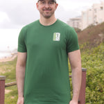 A tall and slim man standing by the sea. The smiling model is wearing a green extra long graphic t-shirt with the text tall club and a tree logo. It features a 3" longer body, 100% organic cotton, and is soft & preshrunk. This extra long tall graphic tee is ideal for tall slim men 6'2"+