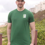 A tall and slim man standing by the sea. The smiling model is wearing a green extra long graphic t-shirt with the text tall club and a tree logo. It features a 3" longer body, 100% organic cotton, and is soft & preshrunk. This extra long tall graphic tee is ideal for tall slim men 6'2"+