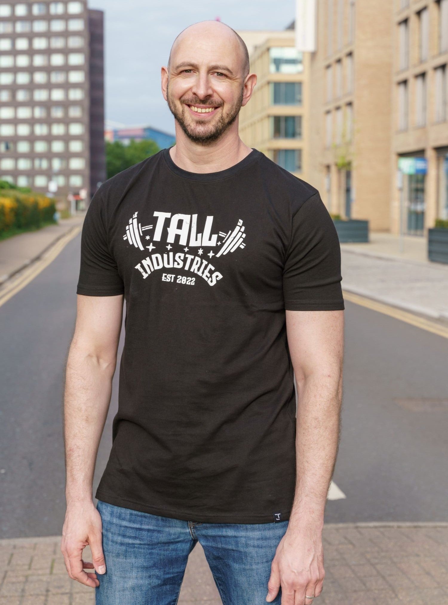 A tall and slim man standing on a pedestrian crossing in the middle of the street. The smiling model is wearing a black extra long graphic t-shirt with the text tall industries and a barbell behind it. It features a 3" longer body, 100% organic cotton, and is soft & preshrunk. This extra long tall graphic tee is ideal for tall slim men 6'2"+.