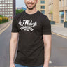 A tall and slim man standing on a pedestrian crossing in the middle of the street. The smiling model is wearing a black extra long graphic t-shirt with the text tall industries and a barbell behind it. It features a 3" longer body, 100% organic cotton, and is soft & preshrunk. This extra long tall graphic tee is ideal for tall slim men 6'2"+.