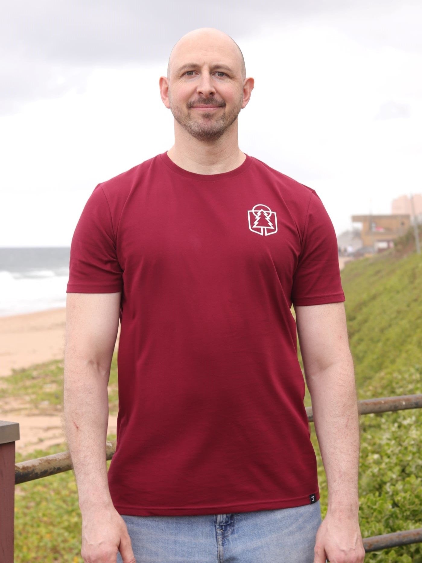 A tall and slim man standing by the sea. The smiling model is wearing a maroon extra long graphic t-shirt with a minimal tree line art design. It features a 3" longer body, 100% organic cotton, and is soft & preshrunk. This extra long tall graphic tee is ideal for tall slim men 6'2"+