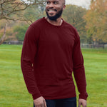An upper body shot of a tall athletic guy wearing an XL tall maroon long sleeve t-shirt, standing in a park and smiling.