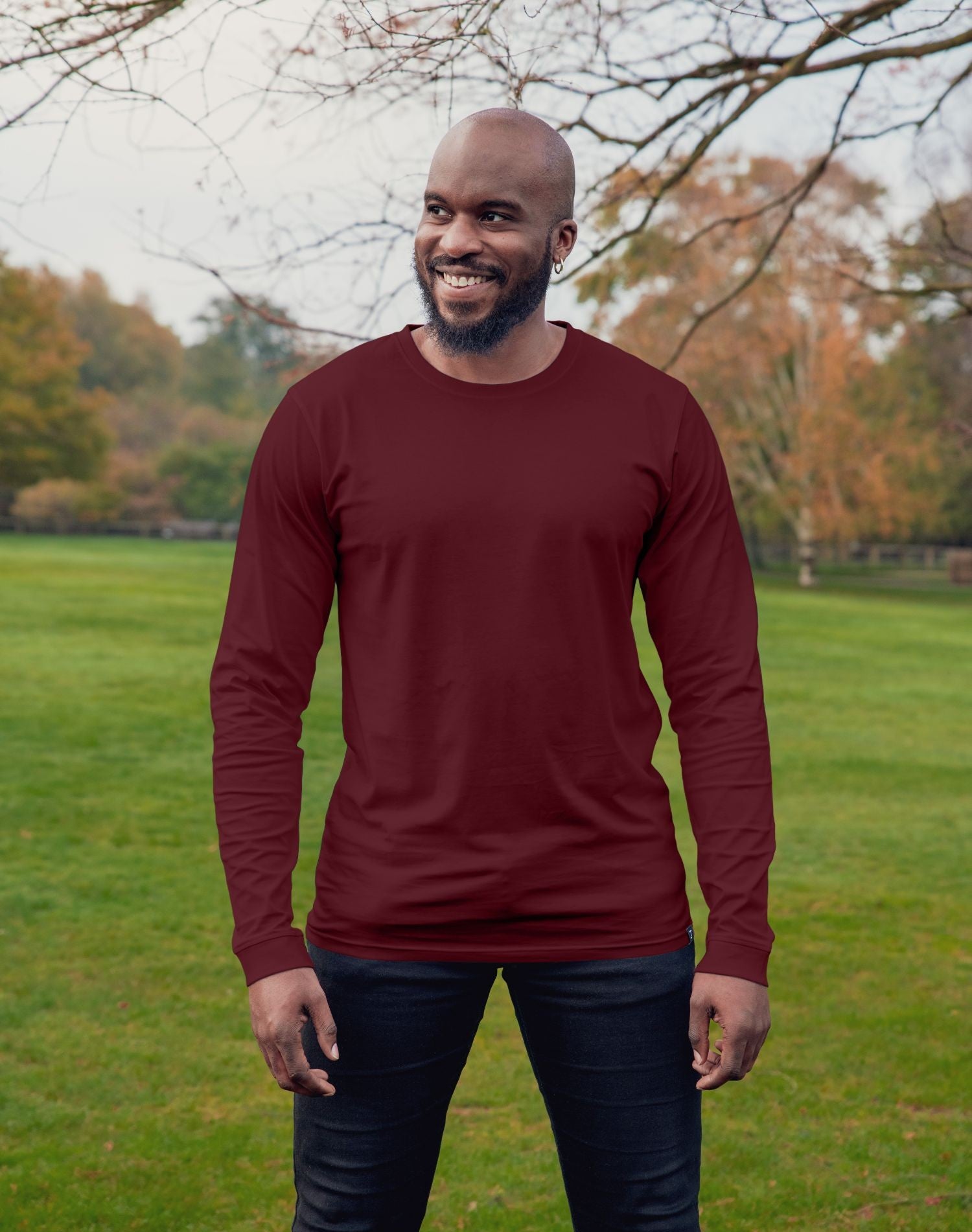An upper body shot of a tall athletic guy wearing an XL tall maroon long sleeve t-shirt, standing in a park and smiling.
