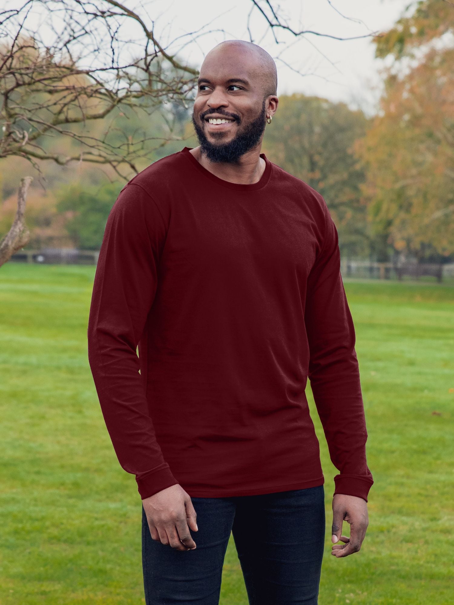 An upper body shot of a tall athletic guy wearing an XL tall maroon long sleeve t-shirt, standing in a park and smiling.