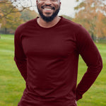 An upper body shot of a tall athletic guy wearing an XL tall maroon long sleeve t-shirt, standing in a park and smiling, hands behind back.