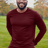 An upper body shot of a tall athletic guy wearing an XL tall maroon long sleeve t-shirt, standing in a park and smiling, hands behind back.