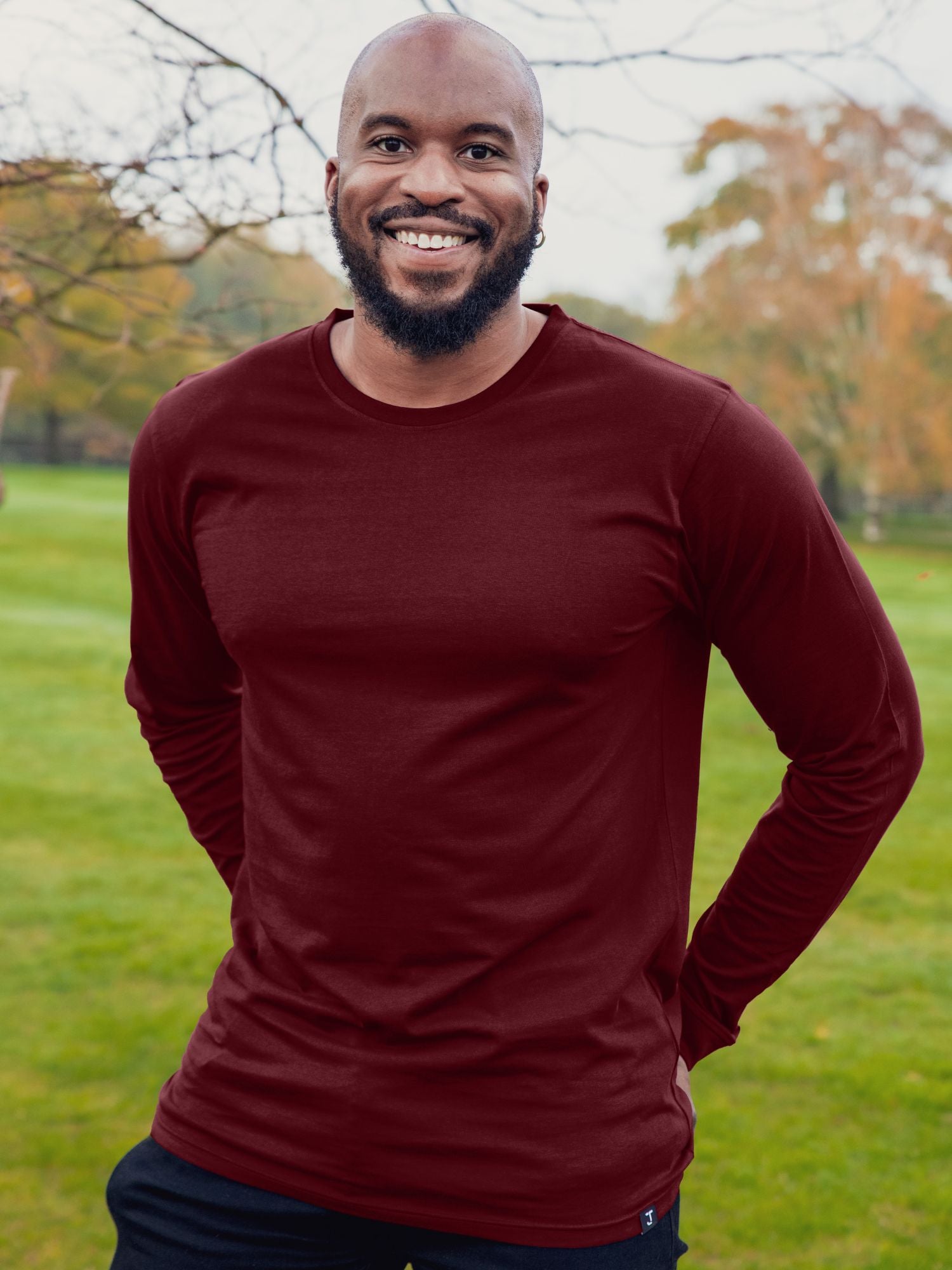 An upper body shot of a tall athletic guy wearing an XL tall maroon long sleeve t-shirt, standing in a park and smiling, hands behind back.
