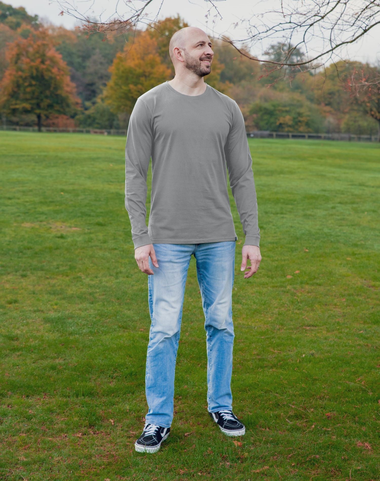 A head to toe shot of a tall athletic guy in a park wearing a light grey long sleeve tall t-shirt, looking to the left.