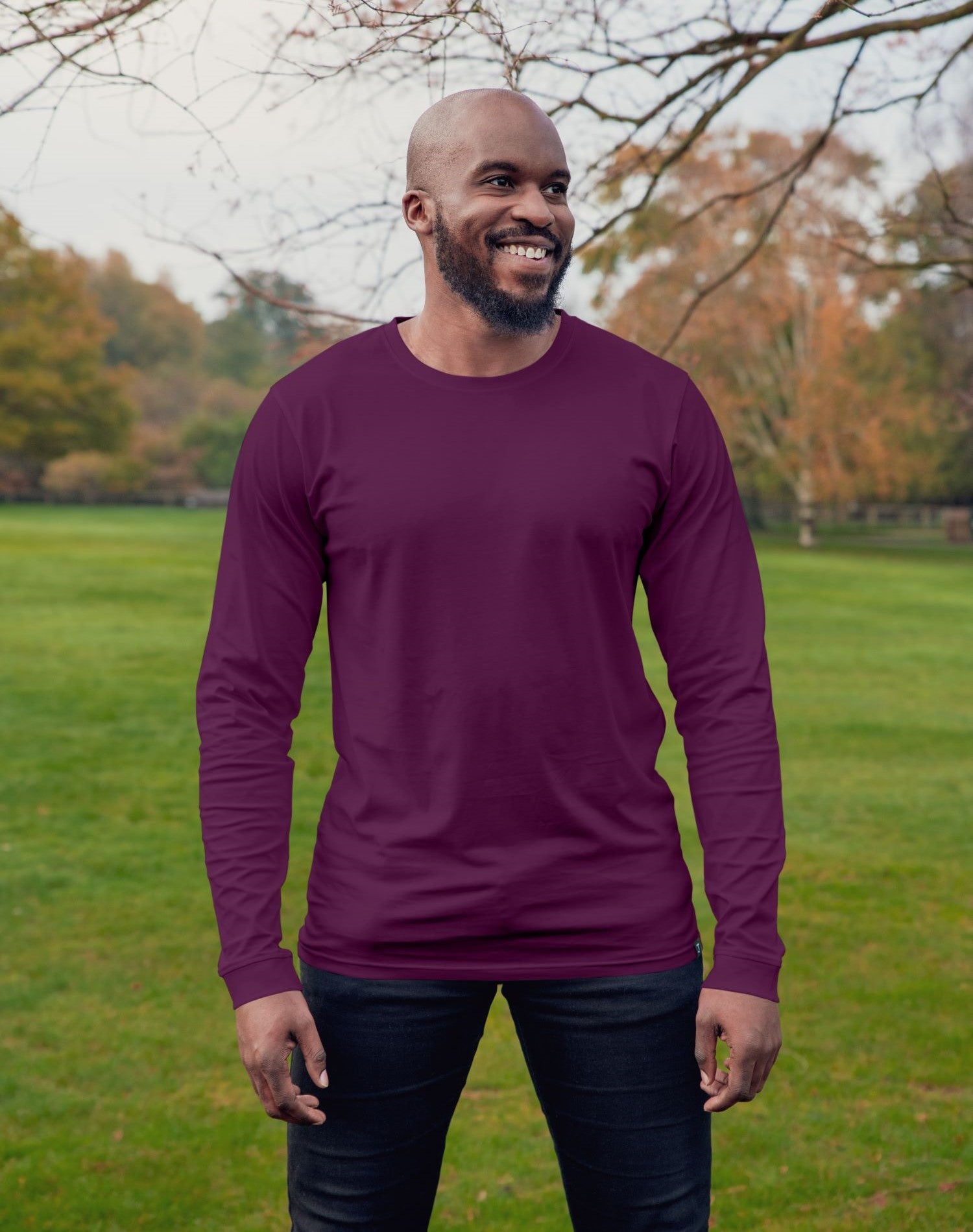 A tall athletic guy wearing a long sleeve dark purple tall t-shirt and smiling in a park while looking to the left.