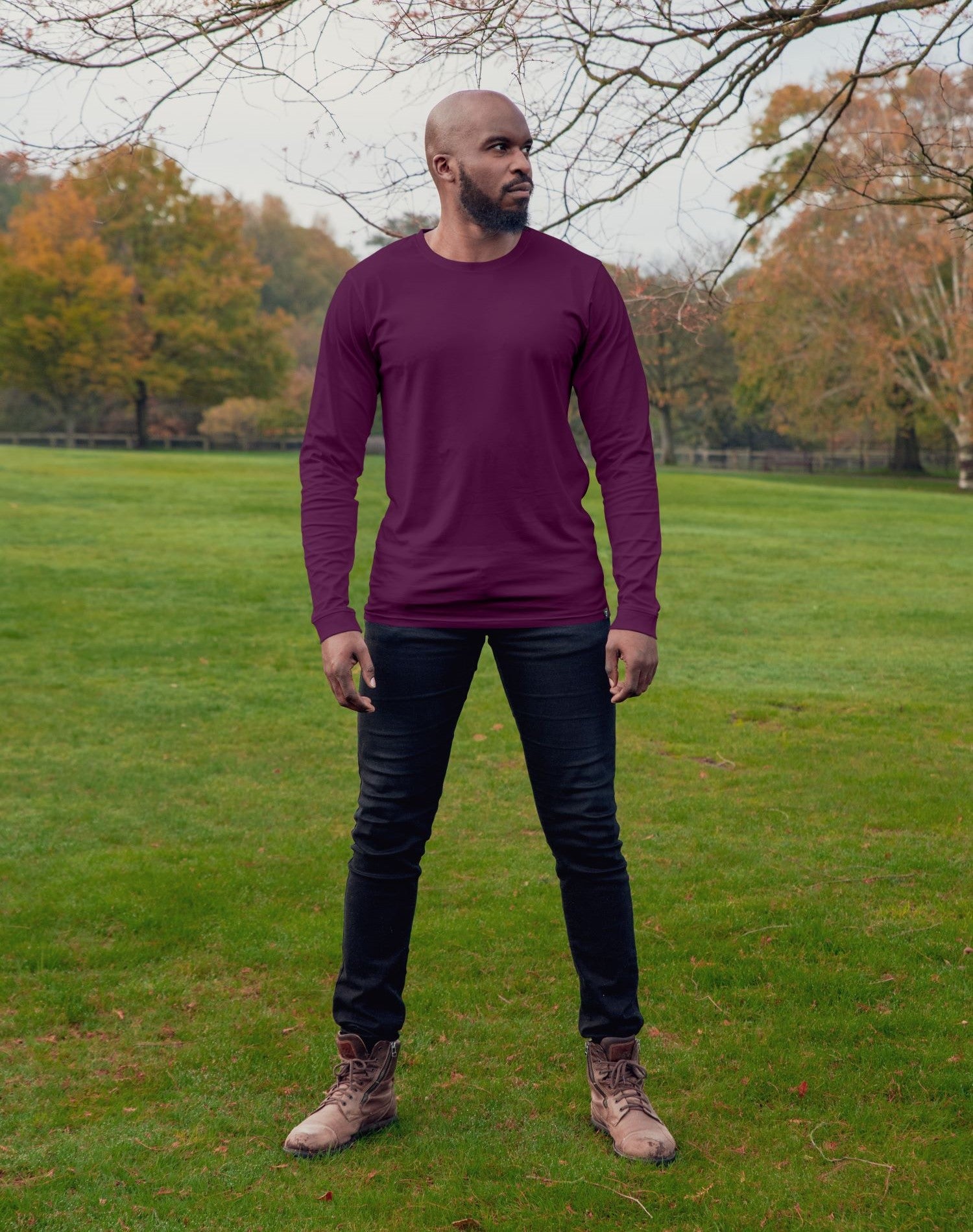 A head to toe shot of a tall lean guy in a  park, wearing a dark purple tall long sleeve t-shirt and looking to the left.