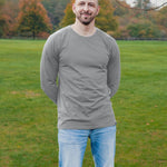 A tall athletic guy wearing a long sleeve light grey tall t-shirt and smiling in a park with hands behind back.