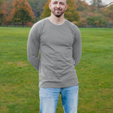 A tall athletic guy wearing a long sleeve light grey tall t-shirt and smiling in a park with hands behind back.