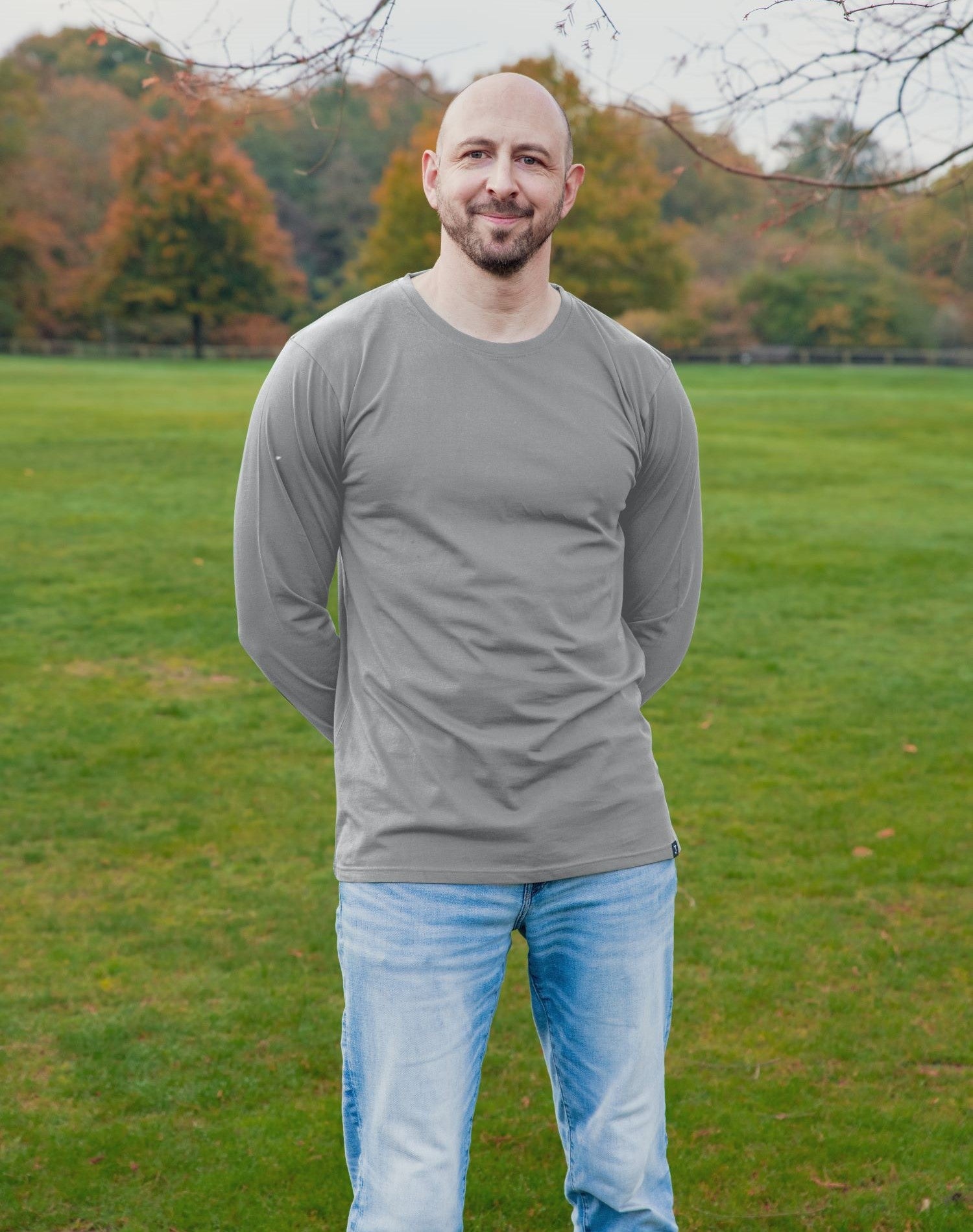 A tall athletic guy wearing a long sleeve light grey tall t-shirt and smiling in a park with hands behind back.