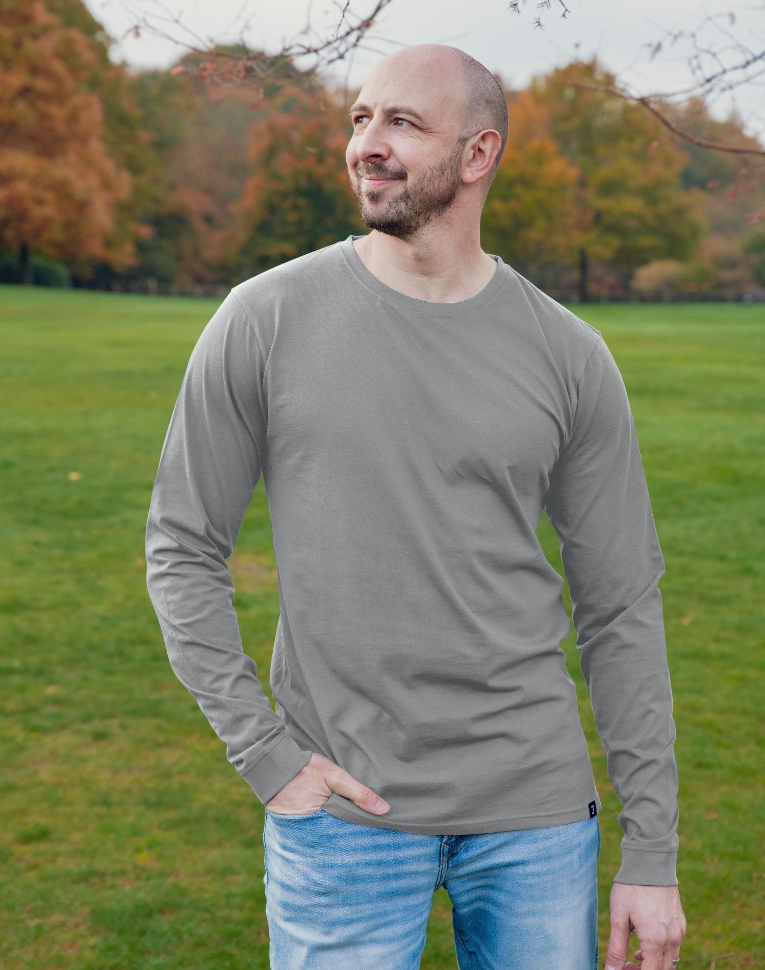 A tall and slim man standing in a park. The smiling model is wearing a light grey long-sleeve tall t-shirt. The tall light grey long-sleeve t-shirt features a 2.5" longer sleeves, 100% organic cotton, and is soft & preshrunk. The light grey long-sleeve t-shirt is ideal for tall slim men 6'2"+.