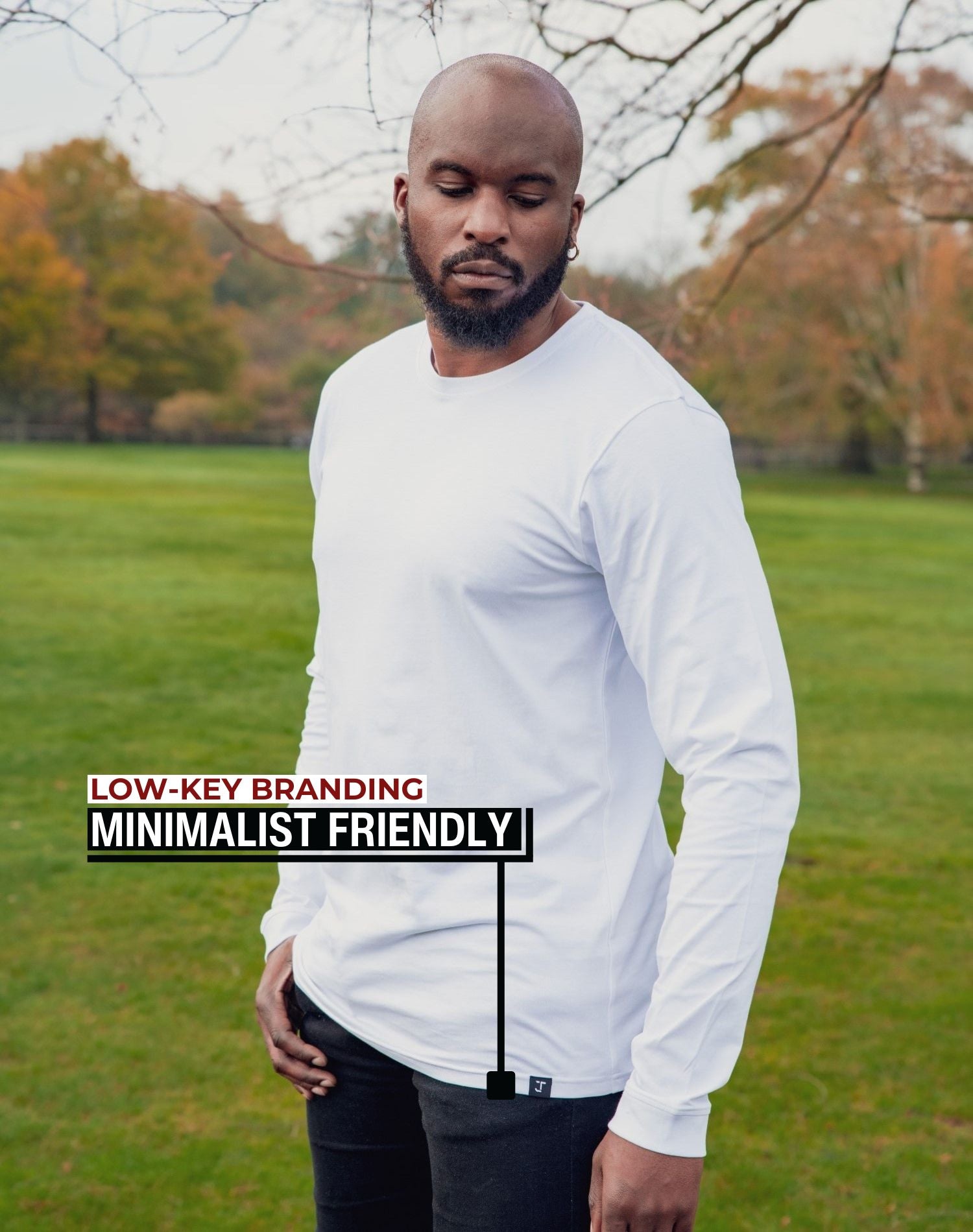 An upper body shot of a tall athletic guy standing in a park wearing a white extra long tall long sleeve t-shirt and looking down at the Just Tall minimalist monogram logo.