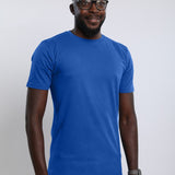 A tall and slim man in the studio standing in front of a light background. The smiling model is wearing an extra long slim medium blue t-shirt in a size large. The tall medium blue t-shirt features a 3" longer body, 100% organic cotton, and is soft & preshrunk. The medium blue t-shirt is ideal for tall slim men 6'2"+.