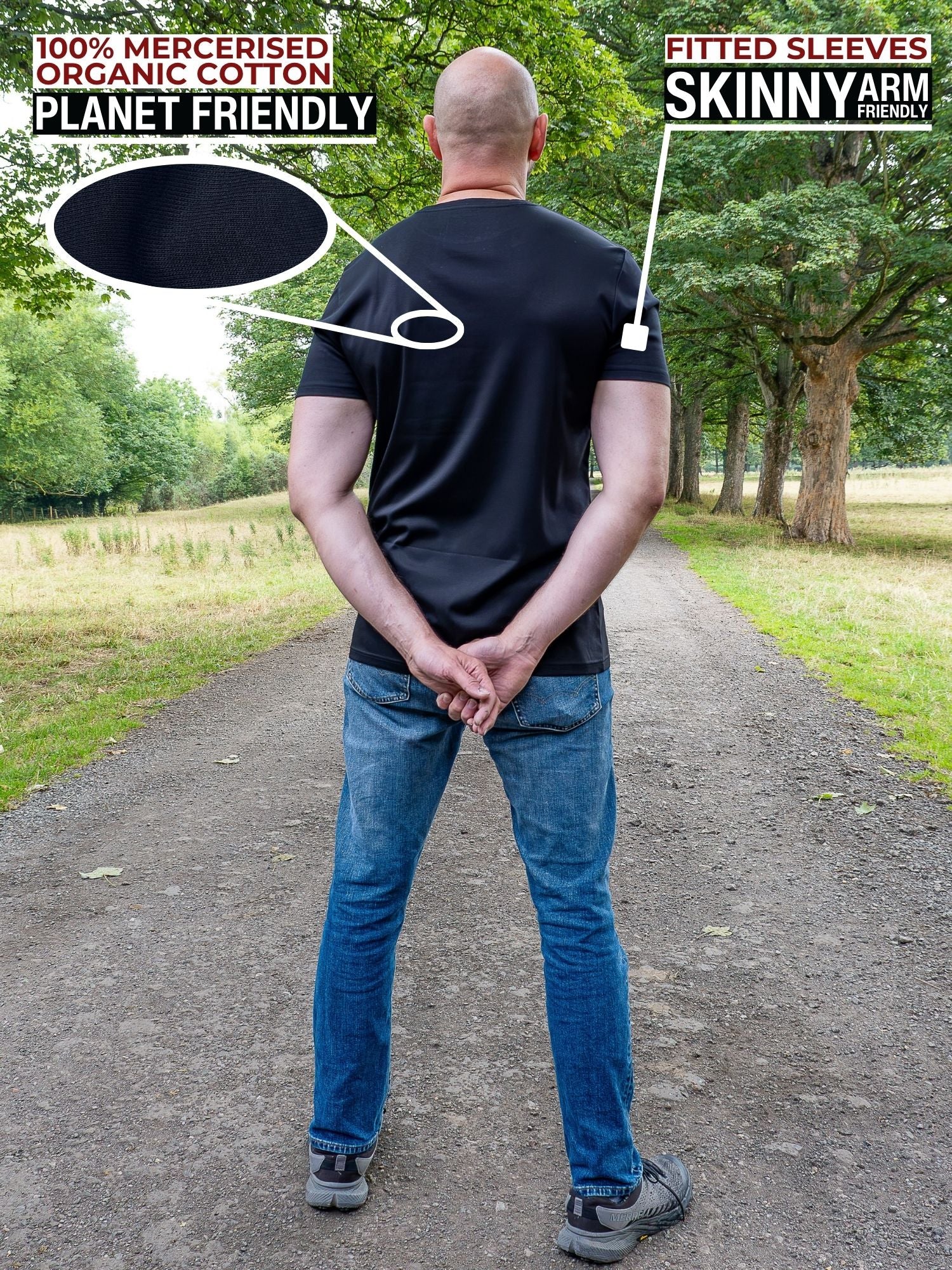 A tall man standing on a park path viewed from behind, wearing a black fitted short-sleeve t-shirt and blue jeans. Text overlays highlight the product features: '100% Mercerised Organic Cotton Planet Friendly' with a fabric swatch circle, and 'Fitted Sleeves Skinny Arm Fit' with a pointer to the sleeve.