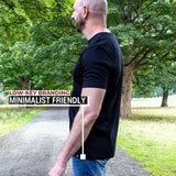 A man wearing a black short-sleeve t-shirt and blue jeans standing on a gravel path in a park with trees. Text on the left reads 'LOW-KEY BRANDING MINIMALIST FRIENDLY' with a white line pointing to the hem wrap logo on the bottom hem.