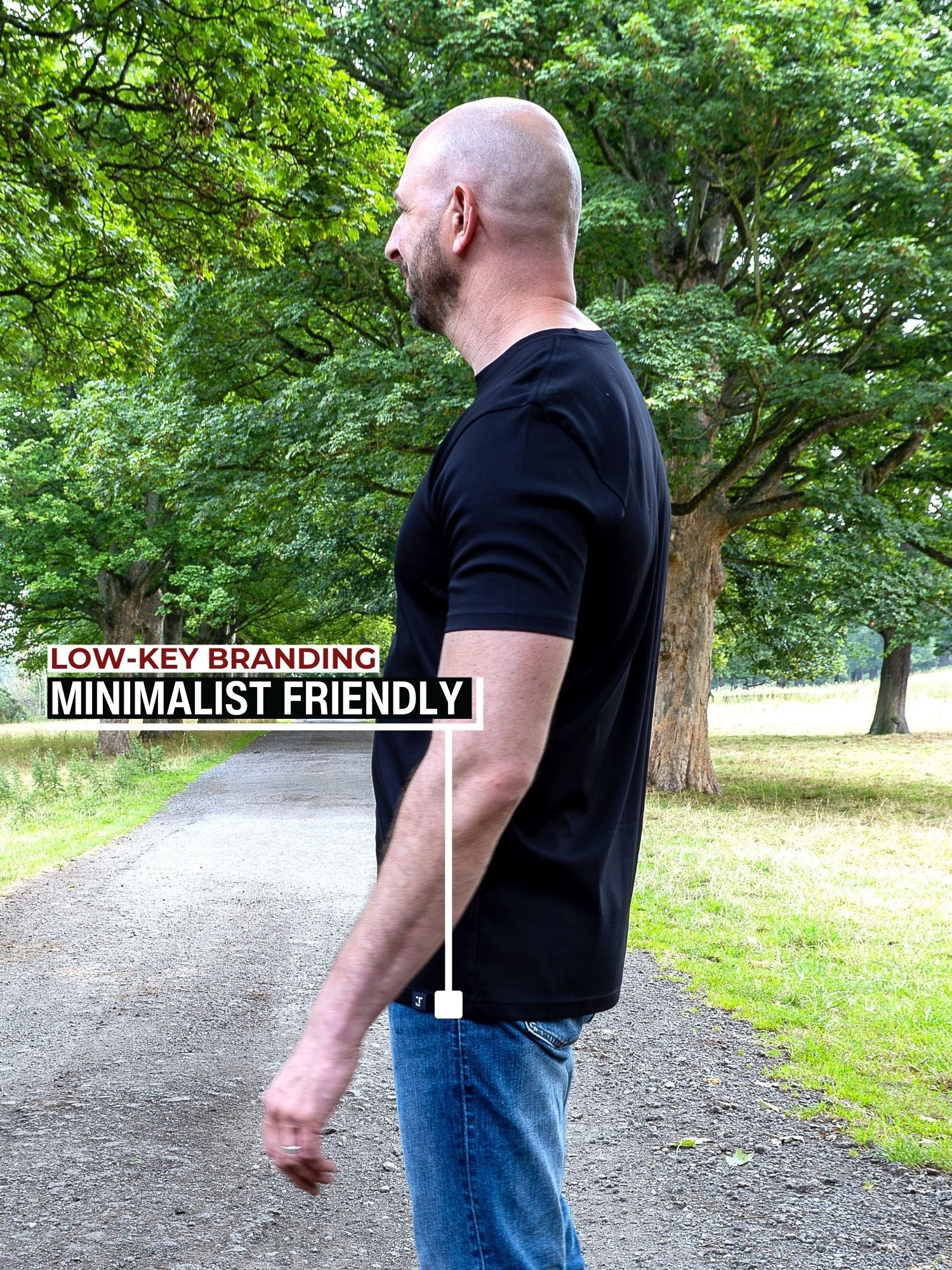 A man wearing a black short-sleeve t-shirt and blue jeans standing on a gravel path in a park with trees. Text on the left reads 'LOW-KEY BRANDING MINIMALIST FRIENDLY' with a white line pointing to the hem wrap logo on the bottom hem.