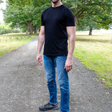 A tall man standing on a gravel path in a park, wearing a black crew-neck t-shirt, blue jeans, and dark gray sneakers.