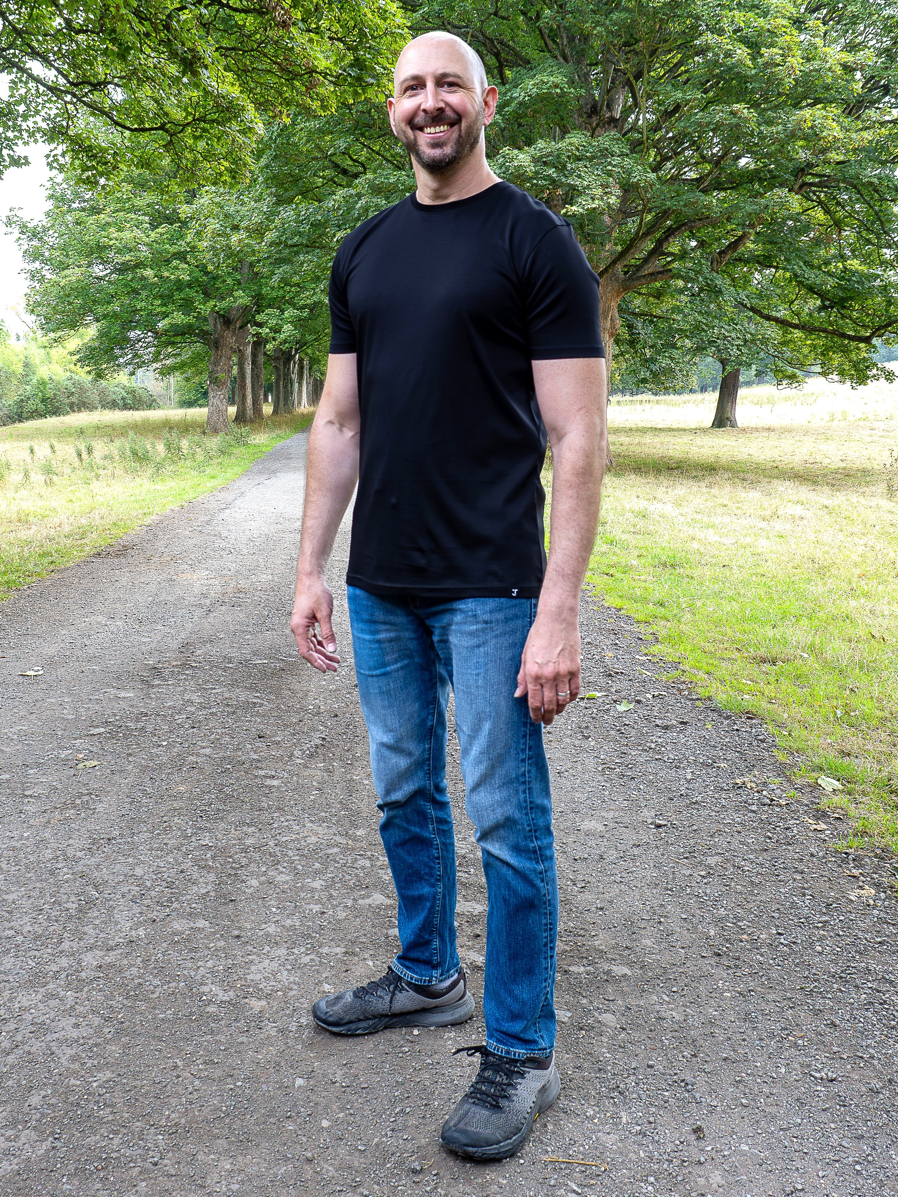 A tall man standing on a gravel path in a park, wearing a black crew-neck t-shirt, blue jeans, and dark gray sneakers.