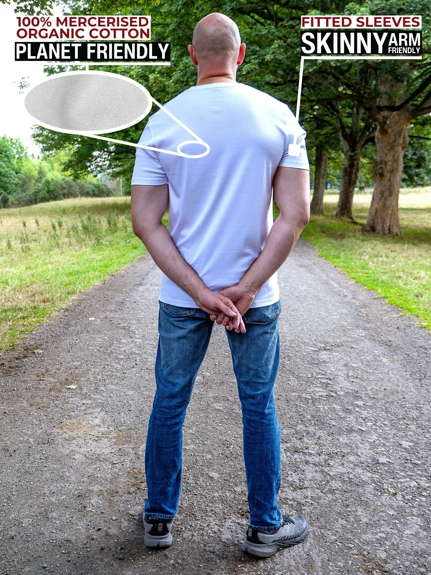 A tall man standing outdoors on a gravel path wearing a white fitted short-sleeve t-shirt and blue jeans, with text overlays highlighting the shirt's features including '100% Mercerised Organic Cotton Planet Friendly' and 'Fitted Sleeves Skinny Arm Fit'.