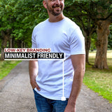 A smiling bald tall man with a beard stands on a tree-lined path wearing a plain white crew-neck t-shirt and blue jeans. Text on the left reads 'LOW-KEY BRANDING MINIMALIST FRIENDLY' with a vertical line pointing to the t-shirt.
