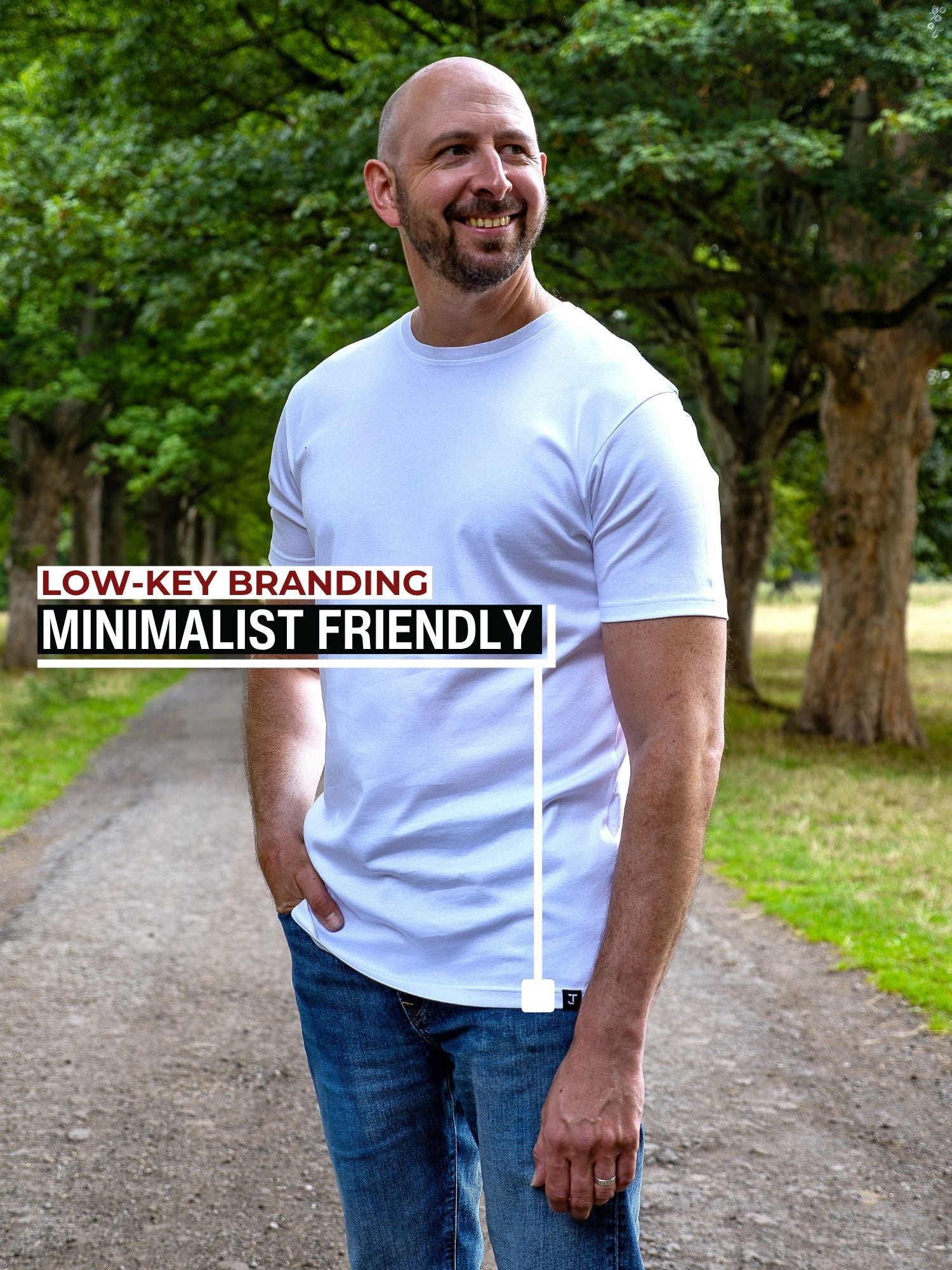A smiling bald tall man with a beard stands on a tree-lined path wearing a plain white crew-neck t-shirt and blue jeans. Text on the left reads 'LOW-KEY BRANDING MINIMALIST FRIENDLY' with a vertical line pointing to the t-shirt.
