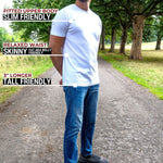 A tall man standing on a park path wearing a white fitted short-sleeve t-shirt and blue jeans, with text callouts describing the shirt's fit and length.