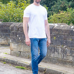 A tall man wearing a plain white crew-neck t-shirt, blue jeans, and sunglasses stands outdoors on a stone path with green trees and bushes in the background.