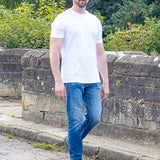 A tall man wearing a plain white crew-neck t-shirt, blue jeans, and sunglasses stands outdoors on a stone path with green trees and bushes in the background.