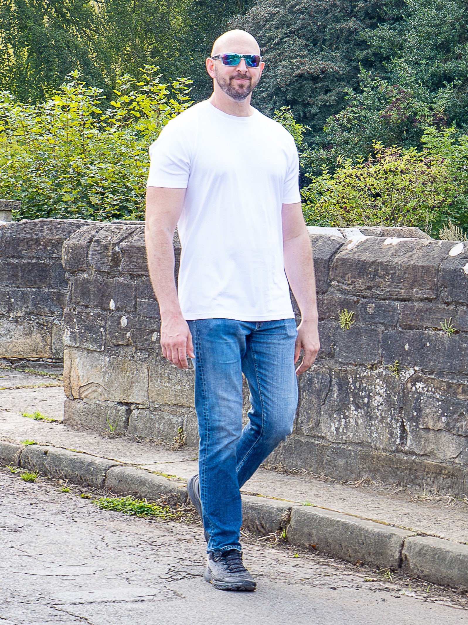 A tall man wearing a plain white crew-neck t-shirt, blue jeans, and sunglasses stands outdoors on a stone path with green trees and bushes in the background.