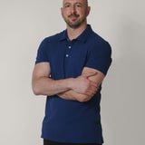 A tall and slim man in the studio standing in front of a light background with his arms crossed. The smiling model is wearing an extra long slim navy blue polo shirt in a size XL. The tall navy blue polo shirt features a 3" longer body, 100% organic cotton, and is soft & preshrunk. The navy blue polo shirt is ideal for tall slim men 6'2"+.